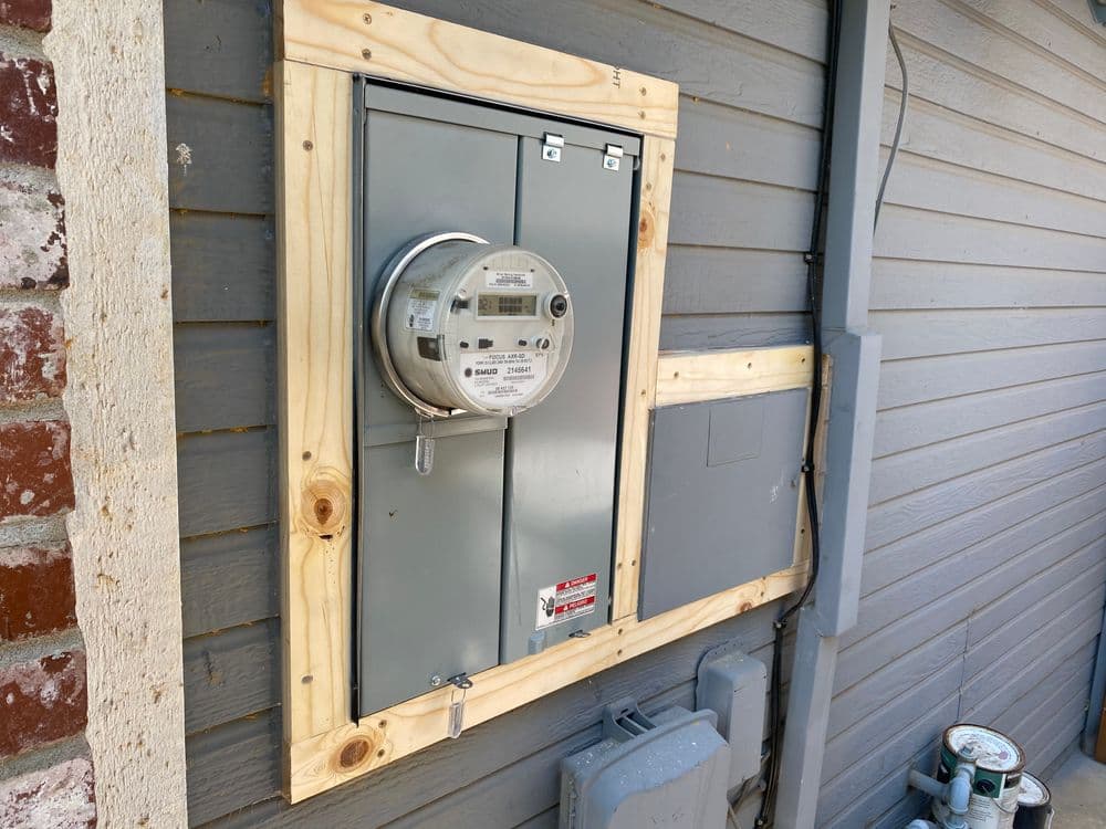 Electric meter mounted on a wooden frame on a gray house exterior.