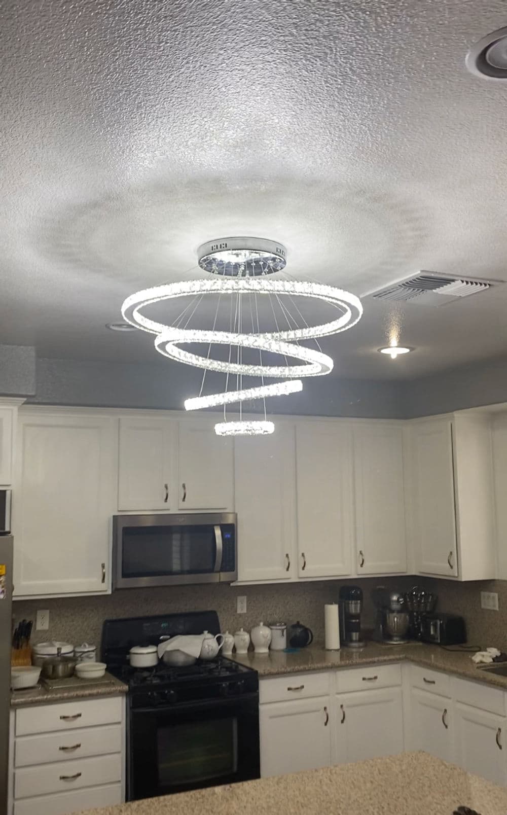 Modern spiral chandelier hanging in a bright kitchen with white cabinets and stainless steel appliances.
