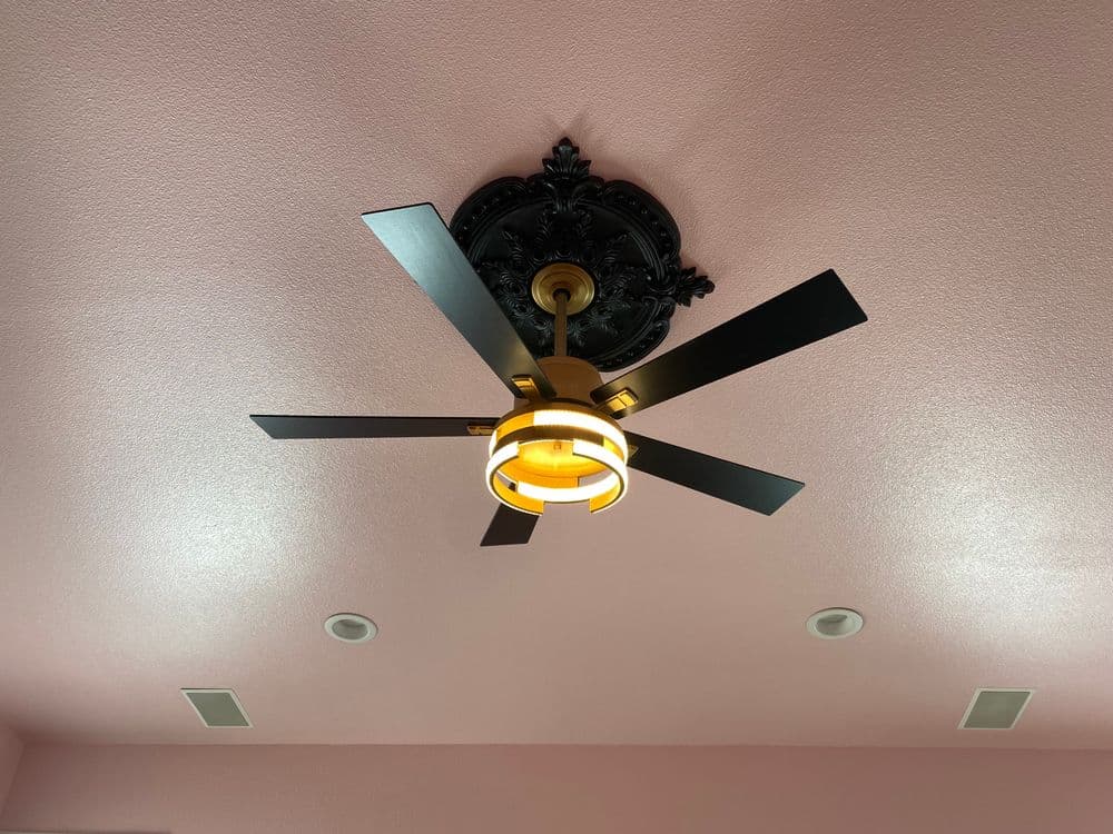 Ceiling fan with black blades and decorative gold light fixture on pink ceiling.