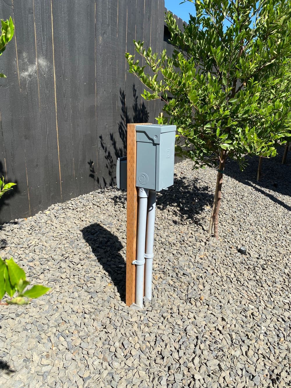 Electrical junction box mounted on wooden post among gravel landscaping and shrubs.