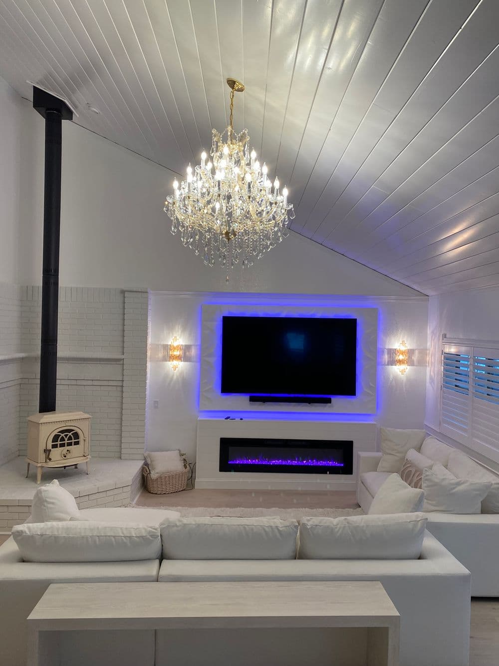 Modern living room with white decor, fireplace, chandelier, and TV surrounded by ambient lighting.